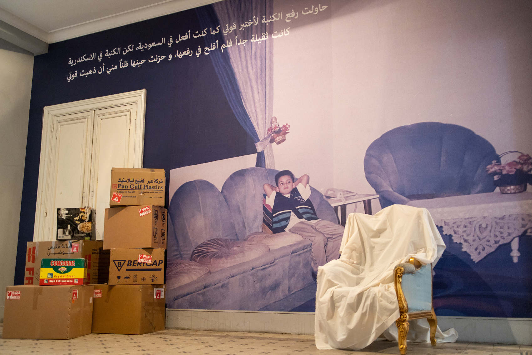 Figure 5: Lina El-Shamy’s installation “Deferred Homes and Boxed Objects: From the Gulf to Egypt.” On the wall, in Arabic: “I tried to lift the sofa to test my strength as I used to do in Saudi Arabia, but the sofa in Alexandria was too heavy and I was not able to lift it, and I felt sad, then, thinking that my strength was gone.”